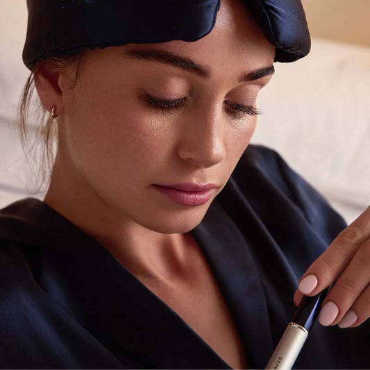 Revitalash Drowsy Collab Collection Image of model with long eyelashed just after waking up with product in her hand and eye mask resting on her head the eye mask is navy silk
