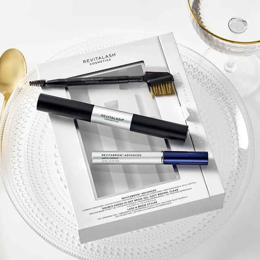 Revitalash Cosmetics brow grooming kit with three tools (spoolie, brush, and brow lamination wand) arranged on their white product box, styled on a white textured plate with a gold spoon and a gold-rimmed glass.