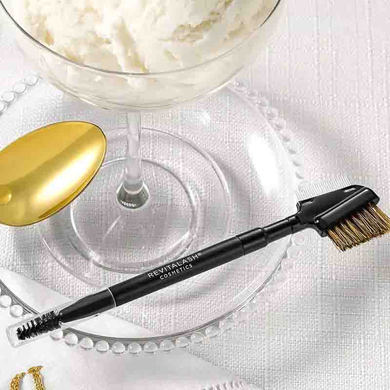 A black RevitaSkin cosmetic brow spoolie and brow brush set arranged on a clear glass plate with a gold spoon and a glass bowl of whipped cream in the background.