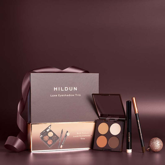 Hildun Luxe Eyeshadow Trio gift set with a dark brown presentation box, a matching inner box, an open four-color eyeshadow palette, a black eyeliner pencil, and a brown eyebrow pencil on a dark brown background.