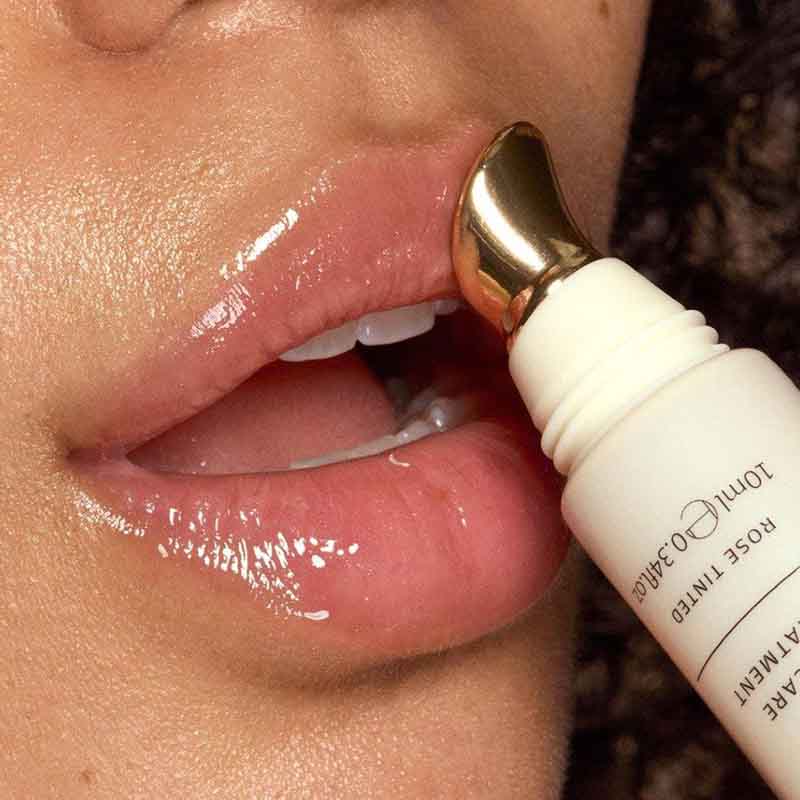 Close-up of a person's lips using a lip treatment wand with a gold applicator tip on a white tube labeled ROSE TINTED LIP CARE.