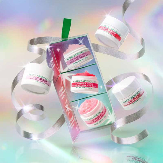 Mario Badescu skincare gift set featuring three white jars of lip balm and exfoliating scrub arranged around a clear gift box with silver ribbons and pastel rainbow lighting effects.