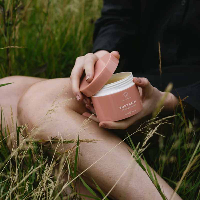 Skin Formulas Body Balm main image with a white background in a woman's hands, half open whilst she sits in a meadow