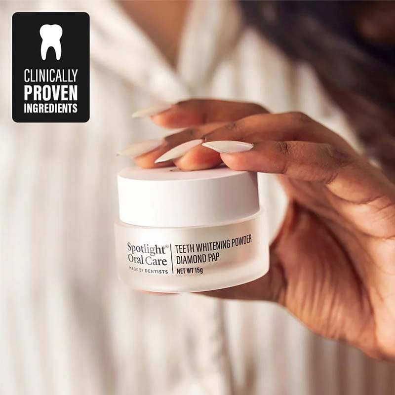 A hand with long nude manicured nails holds a small white jar of Spotlight Oral Care Teeth Whitening Powder against a soft-focus background of a white robe. A black badge in the top-left corner reads 'Clinically Proven Ingredients'.