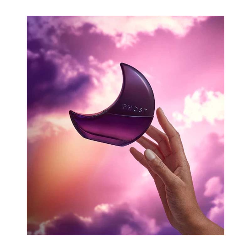 A hand holds a crescent moon-shaped purple perfume bottle labeled 'GHOST' against a dramatic pink and purple cloudy sky background.