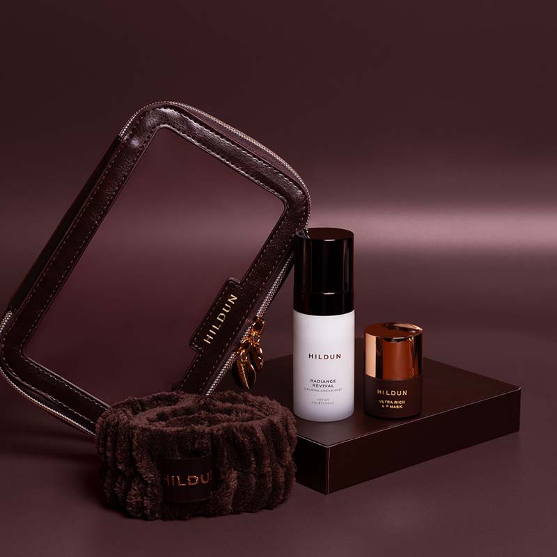 MILDUN skincare set featuring a dark brown faux-leather makeup bag, a matching brown plush neck warmer, a white bottle of Radiance Serum, and a small brown jar with a metallic copper lid, arranged on a dark brown platform against a dark plum background.