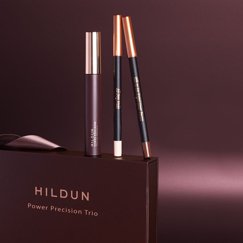 Hildun Power Precision Trio makeup set displayed on a dark brown box, featuring a dark purple tube with a rose gold cap, a black and rose gold pencil, and a black pencil with a white tip, against a dark brown background.