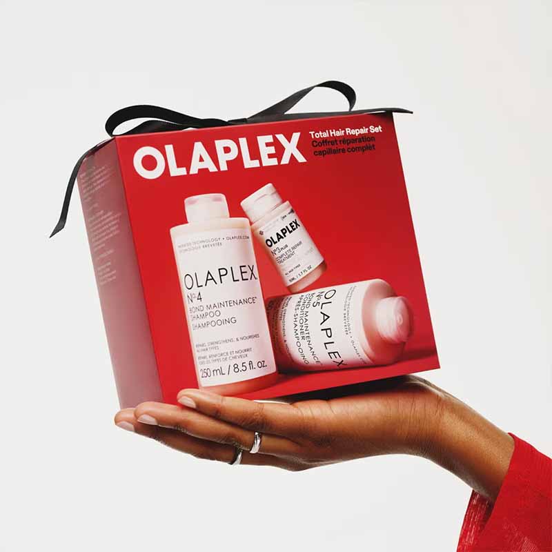 A hand holds an Olaplex K-4 Total Hair Repair Set gift box, which is red with a black ribbon and features images of the shampoo and conditioner bottles on the front, against a plain white background.
