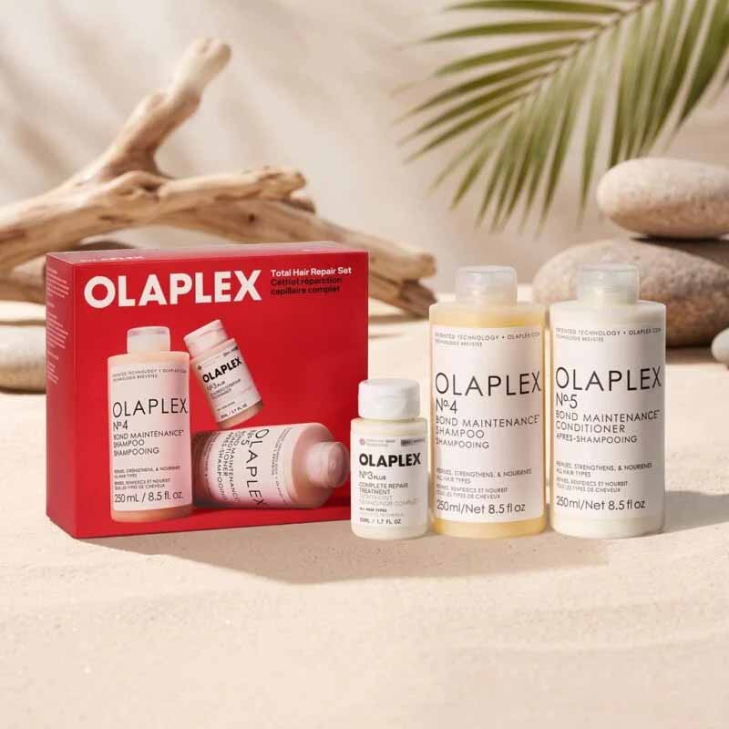 Olaplex Total Hair Repair Set containing a red retail box and four white bottles of shampoo, conditioner, and treatment arranged on a beige surface with driftwood and palm leaves in the background.