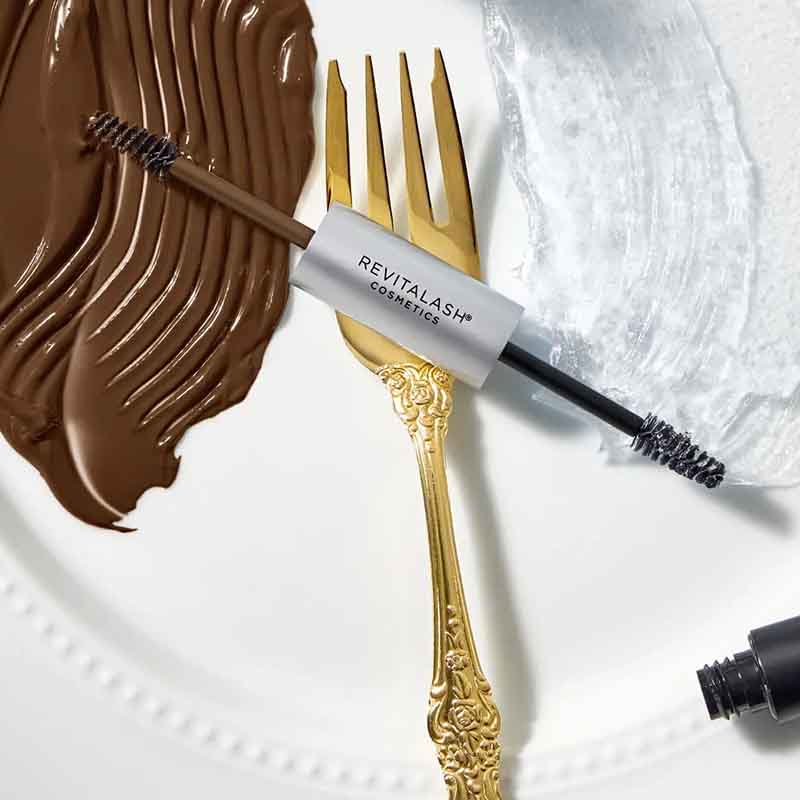 Revitalash Cosmetics brow mascara with a brown applicator wand resting on a gold fork over a white plate with brown and white cream makeup swatches.