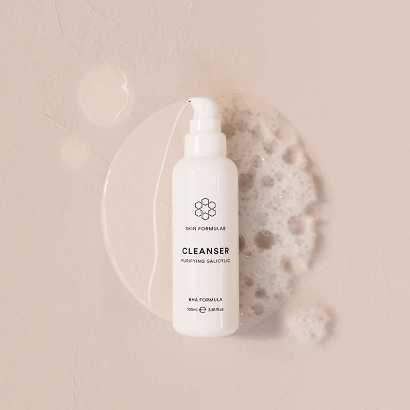 A white bottle of Skin Formulas Cleanser with a pump dispenser, styled on a textured beige background with a clear liquid droplet and a piece of white foam.