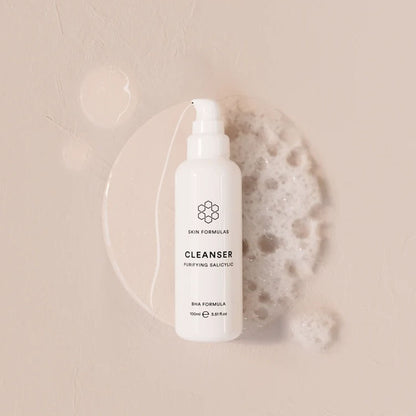 A white bottle of Skin Formulas Cleanser with a pump dispenser, styled on a textured beige background with a clear liquid droplet and a piece of white foam.