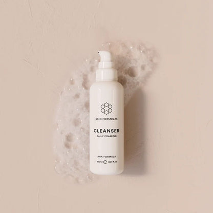 A white pump bottle of Skin Formulas Cleanser Daily Foaming cleanser, surrounded by white foam bubbles on a textured beige background.