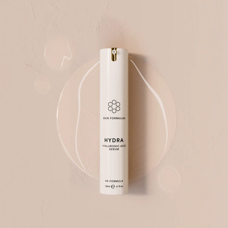 A white cylindrical skincare pump bottle labeled 'HYDRA HYALURONIC ACID SERUM' by Skin Formulas, set against a warm beige textured background with a clear circular gel smear behind it.