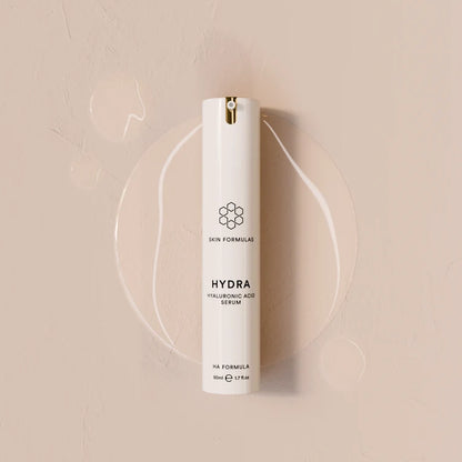 A white cylindrical skincare pump bottle labeled 'HYDRA HYALURONIC ACID SERUM' by Skin Formulas, set against a warm beige textured background with a clear circular gel smear behind it.