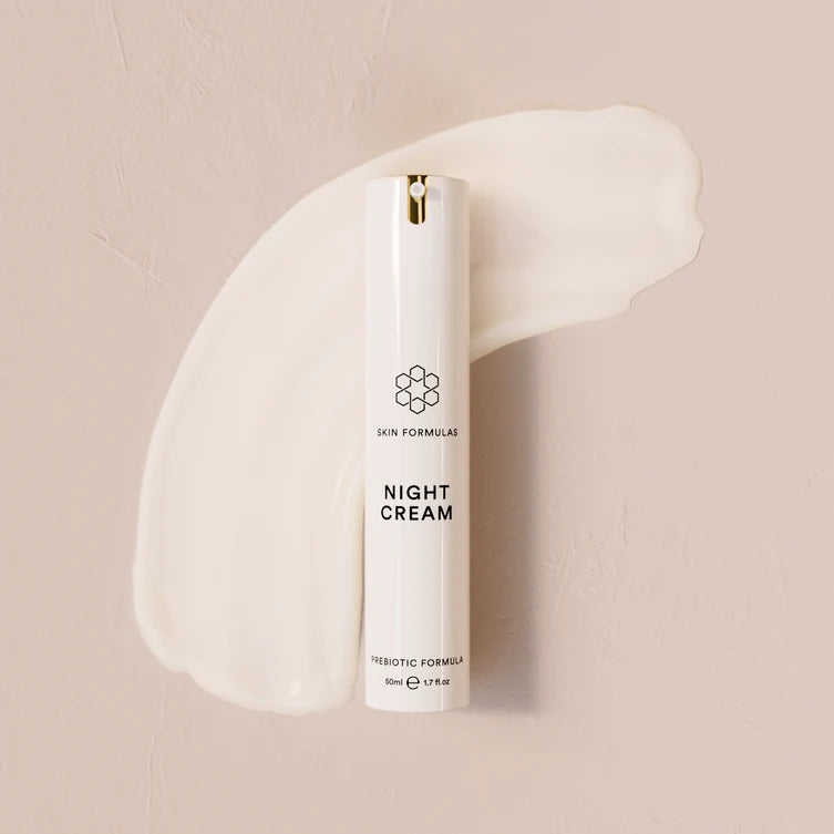 A white cosmetic tube labeled 'SKIN FORMULAS NIGHT CREAM' stands upright against a warm beige textured background with a cream-colored cream smear behind it.