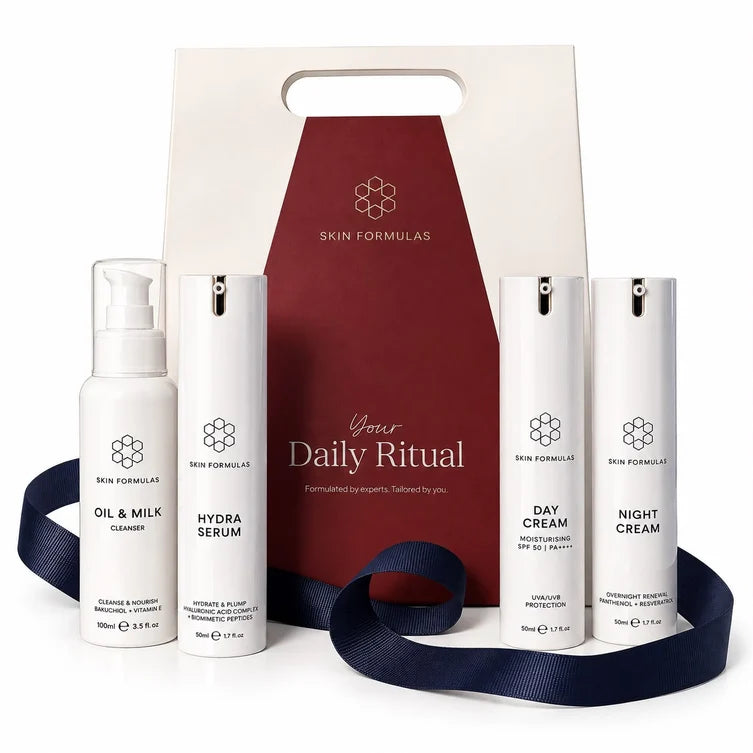 A skincare gift set from Skin Formulas featuring five white bottles (Oil & Milk Cleanser, Hydra Serum, Day Cream, Night Cream, and an inner box) arranged in front of a cream and burgundy gift bag with a navy blue ribbon.