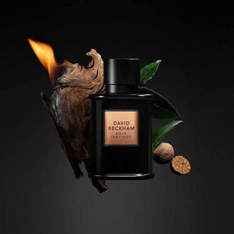 A black David Beckham Elixir Instinct perfume bottle with a gold label, styled with a burning piece of wood, green leaves, and two brown botanical pods on a dark background.