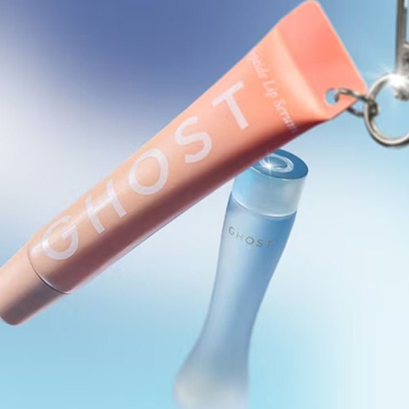 A GHOST-branded beauty set on a light blue gradient background, featuring a square dark blue case with a blue ribbon, a frosted bottle, and an orange tube with a silver keychain.