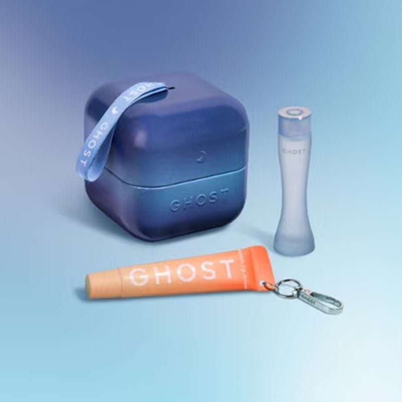A GHOST-branded beauty set on a light blue gradient background, featuring a square dark blue case with a blue ribbon, a frosted bottle, and an orange tube with a silver keychain.