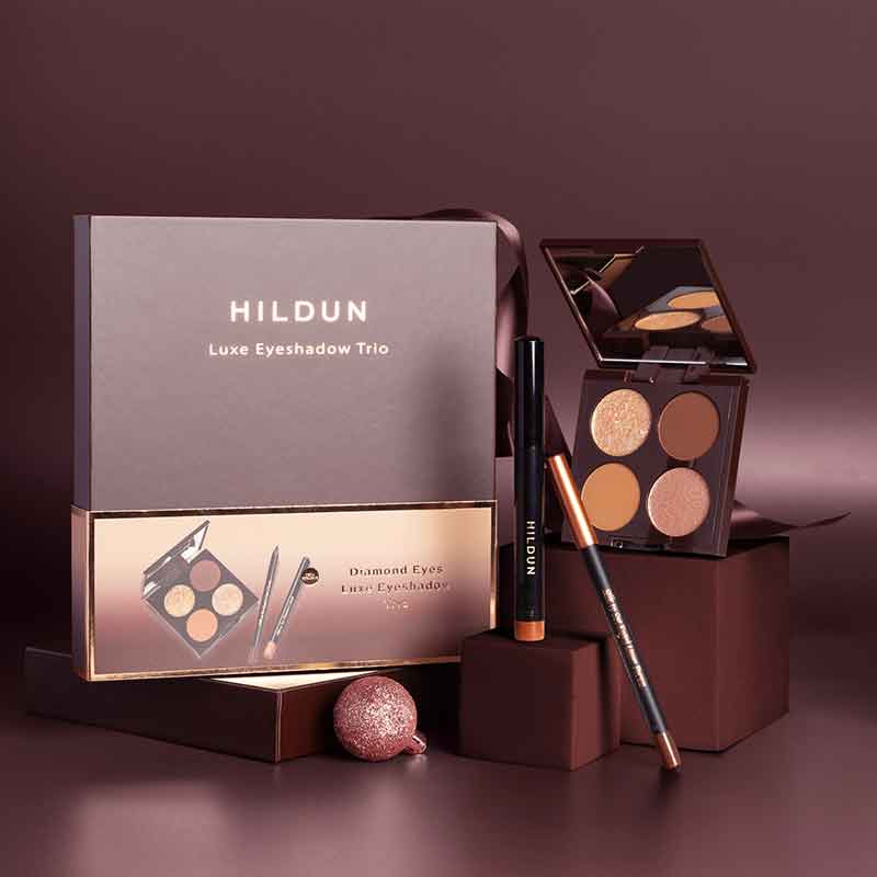Hildun Luxe Eyeshadow Trio makeup set displayed with its outer box, an open eyeshadow palette, a black pencil, and a metallic pencil on dark brown blocks against a mauve-brown studio background.