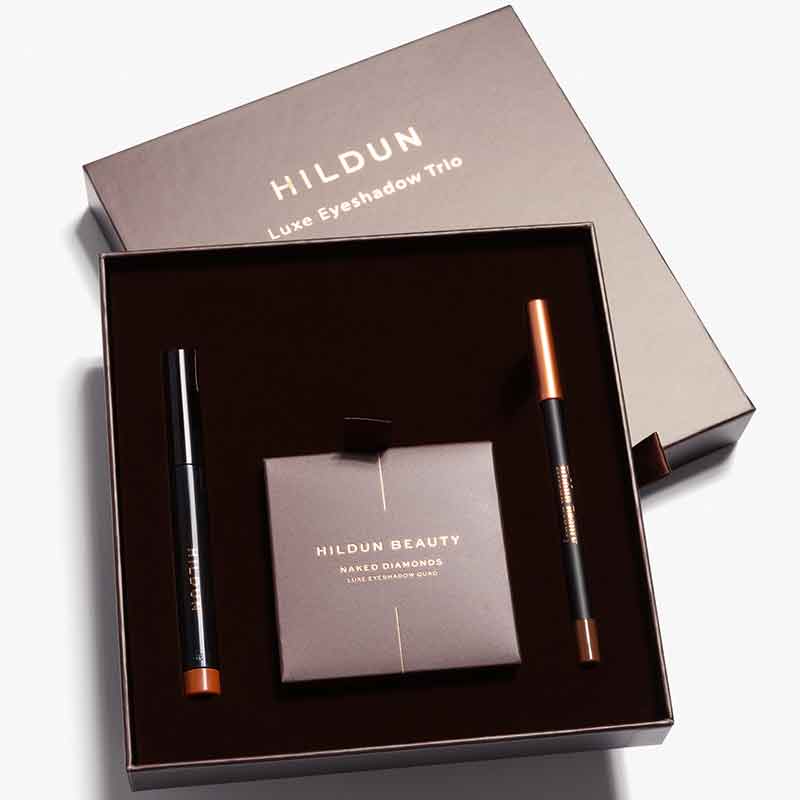 Hildun Luxe Eyeshadow Trio gift set containing a black eyeshadow pencil with copper cap, a matching pencil, and a Hildun Beauty Naked Diamond makeup sponge, presented in a dark brown box.