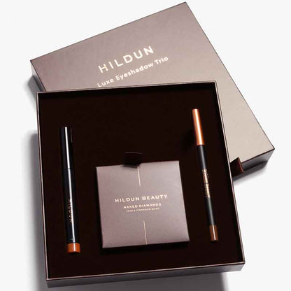 Hildun Luxe Eyeshadow Trio gift set containing a black eyeshadow pencil with copper cap, a matching pencil, and a Hildun Beauty Naked Diamond makeup sponge, presented in a dark brown box.