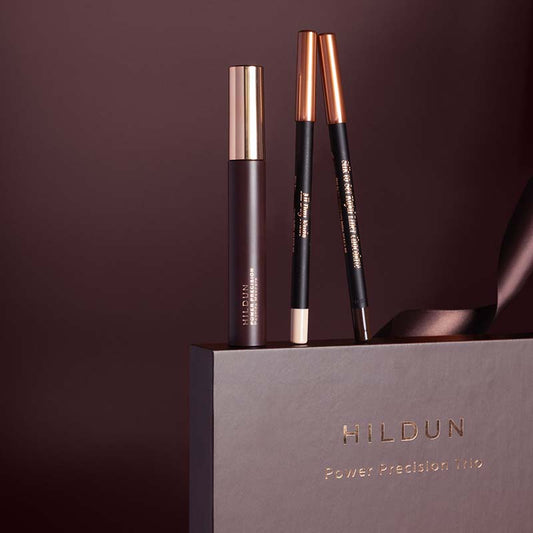 Hildun Power Precision Trio makeup set featuring a dark brown mascara tube and two matching eyebrow pencils with rose gold and black barrels, displayed on top of a dark brown Hildun gift box.