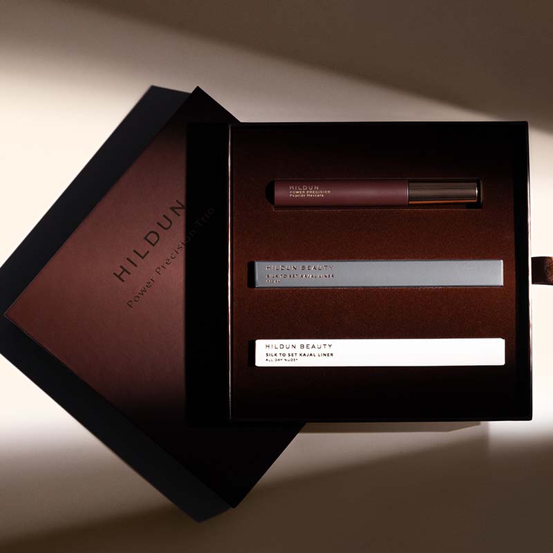 Hildun Beauty Power Translating Set shown in an open dark presentation box, featuring a lip gloss tube, a silver bar, and a white tube on a warm beige background.
