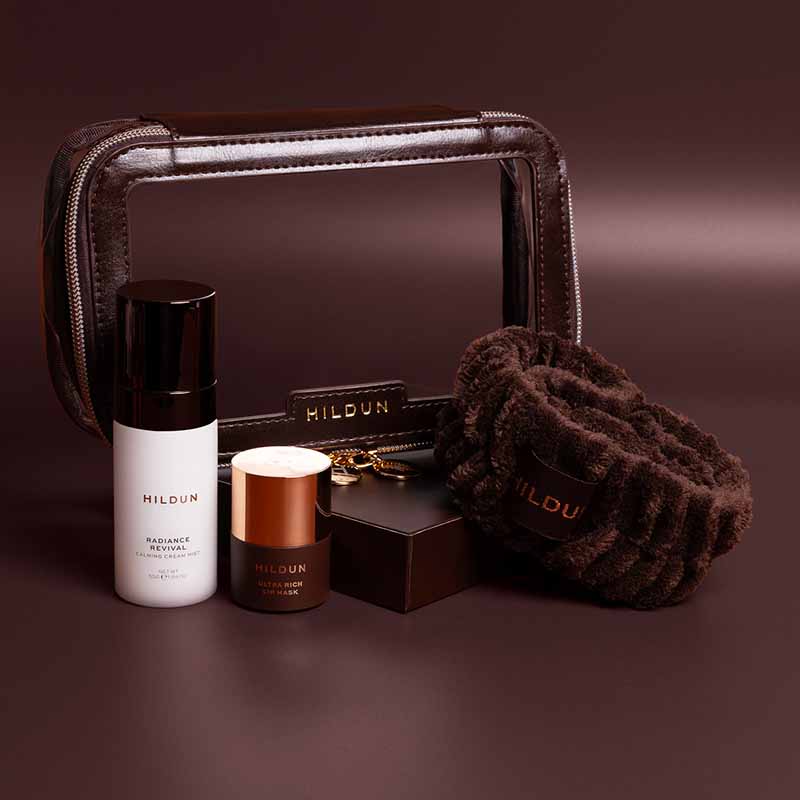 Hildun luxury skincare gift set arranged on a dark brown background, featuring a clear zip cosmetic bag, a white bottle labeled Radiance Reviver, a small jar with a metallic copper lid, a black box, and a brown plush eye mask.