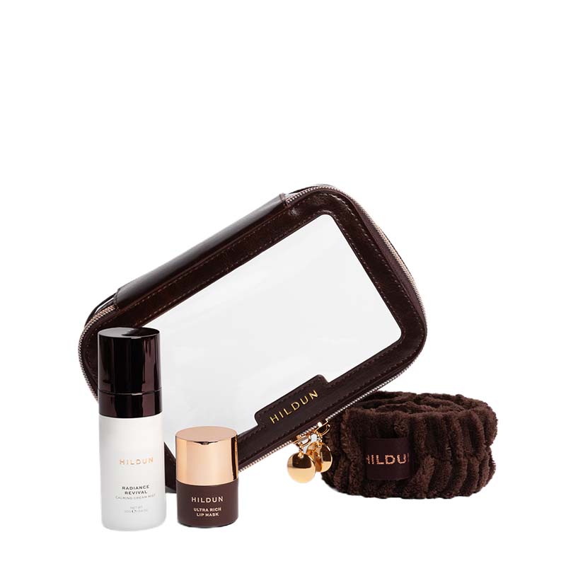 Hildun travel set featuring a brown faux-leather and clear window zippered pouch, a brown hair tie, and two skincare bottles (radiant serum and face primer) on a white background.