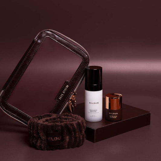 MILDUN skincare set featuring a dark brown faux-leather makeup bag, a matching brown plush neck warmer, a white bottle of Radiance Serum, and a small brown jar with a metallic copper lid, arranged on a dark brown platform against a dark plum background.