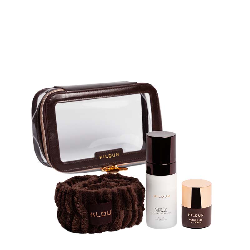 HILDUN skincare set featuring a brown faux-leather makeup bag with a clear window, a brown plush microfiber towel, a white bottle of Radiance Serum, and a small brown jar with a rose gold lid.