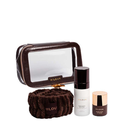HILDUN skincare set featuring a brown faux-leather makeup bag with a clear window, a brown plush microfiber towel, a white bottle of Radiance Serum, and a small brown jar with a rose gold lid.