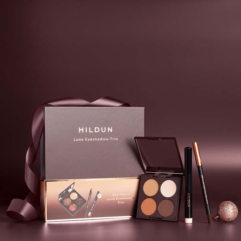Hildun Luxe Eyeshadow Trio gift set with a dark brown presentation box, a matching inner box, an open four-color eyeshadow palette, a black eyeliner pencil, and a brown eyebrow pencil on a dark brown background.