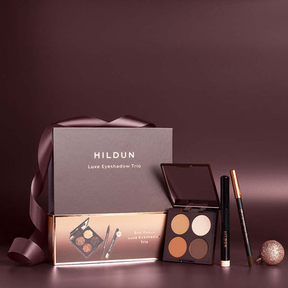 Hildun Luxe Eyeshadow Trio gift set with a dark brown presentation box, a matching inner box, an open four-color eyeshadow palette, a black eyeliner pencil, and a brown eyebrow pencil on a dark brown background.
