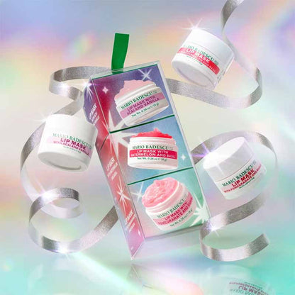 Mario Badescu skincare gift set featuring three white jars of lip balm and exfoliating scrub arranged around a clear gift box with silver ribbons and pastel rainbow lighting effects.