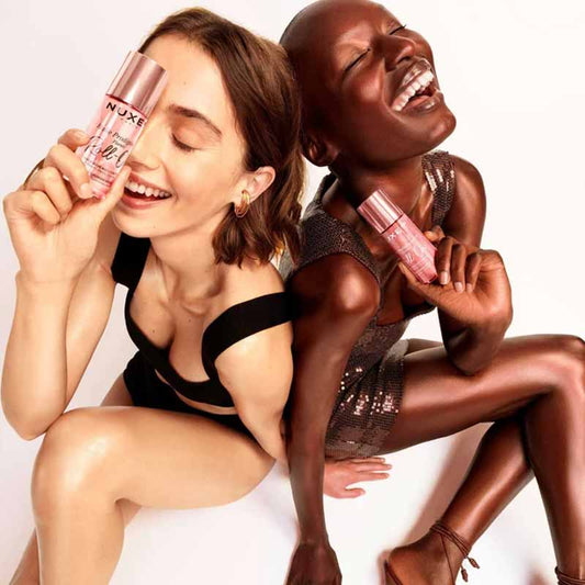 NUXE Huile Prodigieuse Floral Gold Shimmer Multi-Purpose Dry Oil image of two women sitting beside each other in party outfits holding a bottle of the shimmer and the non  shimmer products, their legs and arms are radiant and dewy reflecting the lght