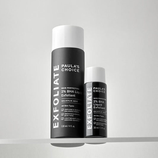 Two skincare bottles from Paula's Choice Exfoliate line, including a larger black canister and a smaller black spray bottle, both with white caps, displayed on a light gray shelf against a neutral background.