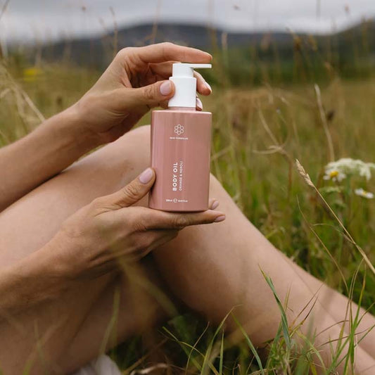 Skin Formulas Body Oil product in a woman's hands, facing the camera with smooth moisturizes legs behind it, the background is in a meadow with long stems of grass and dandelion's blurred behind the legs