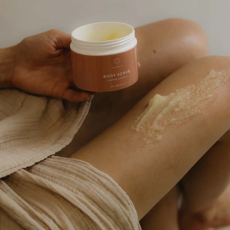 Skin Formulas Body Scrub swatched on a woman's right leg, the product open in her left hand 