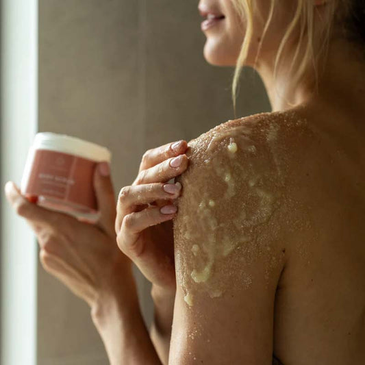 Skin Formulas Body Scrub in a woman's hands, she is using the product on her left shoulder, a hick grainy texture in her hands to smooth and shine