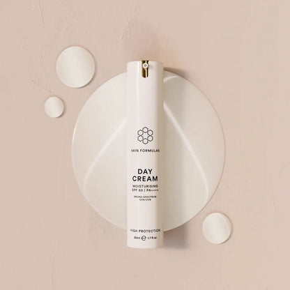 A white cylindrical bottle of SKIN FORMULAS Day Cream SPF 50 PA++++ moisturizer, displayed on a matching white oval plate with scattered white cream droplets on a textured beige background.