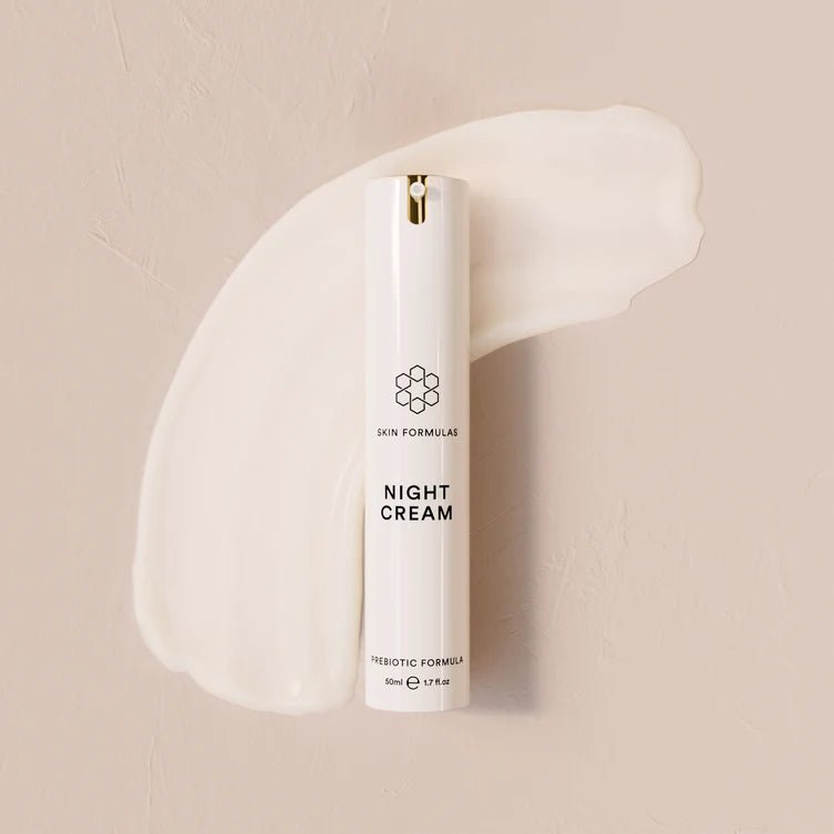 A white cosmetic tube labeled 'SKIN FORMULAS NIGHT CREAM' stands upright against a warm beige textured background with a cream-colored cream smear behind it.