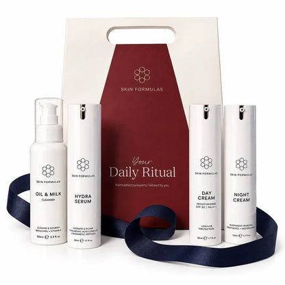 A skincare gift set from Skin Formulas featuring five white bottles (Oil & Milk Cleanser, Hydra Serum, Day Cream, Night Cream, and an inner box) arranged in front of a cream and burgundy gift bag with a navy blue ribbon.