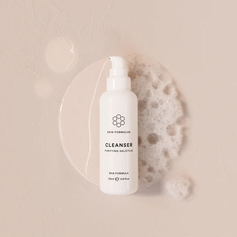 A white bottle of Skin Formulas Cleanser with a pump dispenser, styled on a textured beige background with a clear liquid droplet and a piece of white foam.