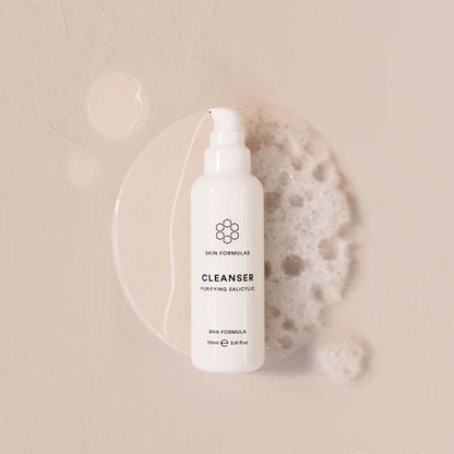 A white bottle of Skin Formulas Cleanser with a pump dispenser, styled on a textured beige background with a clear liquid droplet and a piece of white foam.