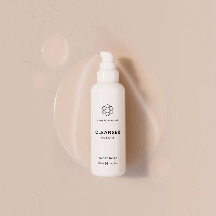 A white pump bottle of Skin Formulas Cleanser Oil & Milk, centered against a warm beige textured background with a soft circular cream smear behind it.