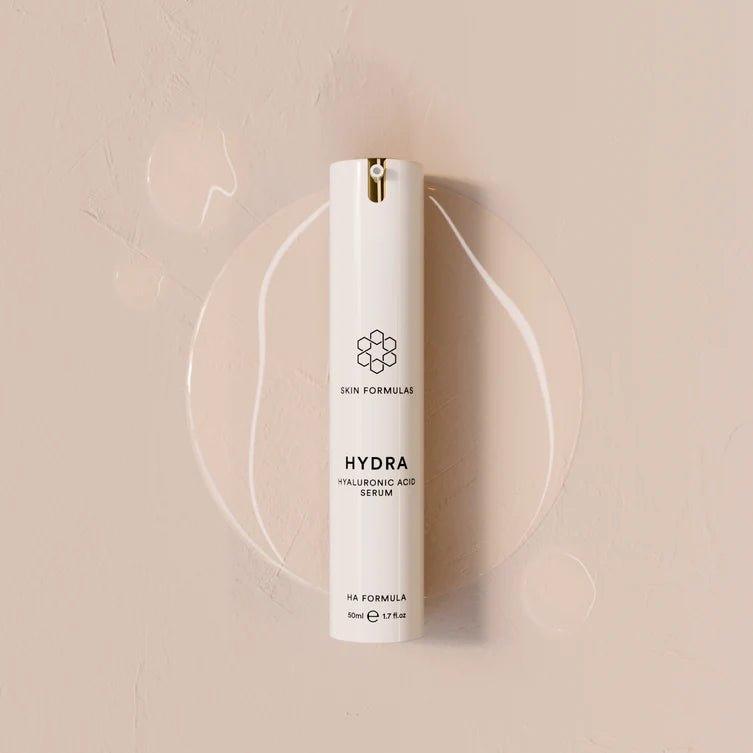 A white cylindrical skincare pump bottle labeled 'HYDRA HYALURONIC ACID SERUM' by Skin Formulas, set against a warm beige textured background with a clear circular gel smear behind it.