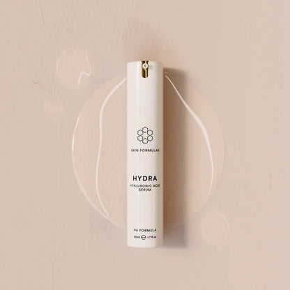 A white cylindrical skincare pump bottle labeled 'HYDRA HYALURONIC ACID SERUM' by Skin Formulas, set against a warm beige textured background with a clear circular gel smear behind it.